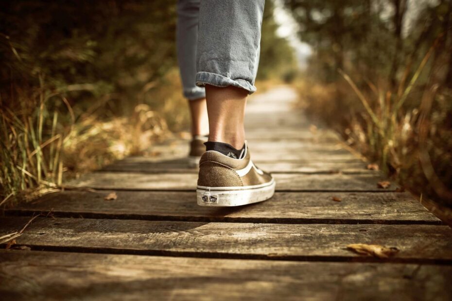 Auf einem Holzsteg, der durch die Natur führt, geht eine Person in Turnschuhen und mit Jeans. Es ist der Weg, die Schuhe, die Waden und das untere Stück der Hosenbeine zu erkennen.