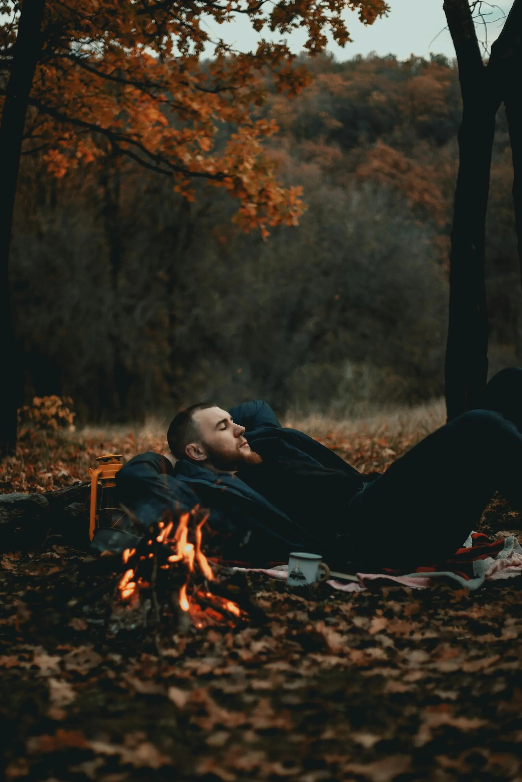 A man relaxes by a campfire in an autumn forest, enjoying the outdoor tranquility.