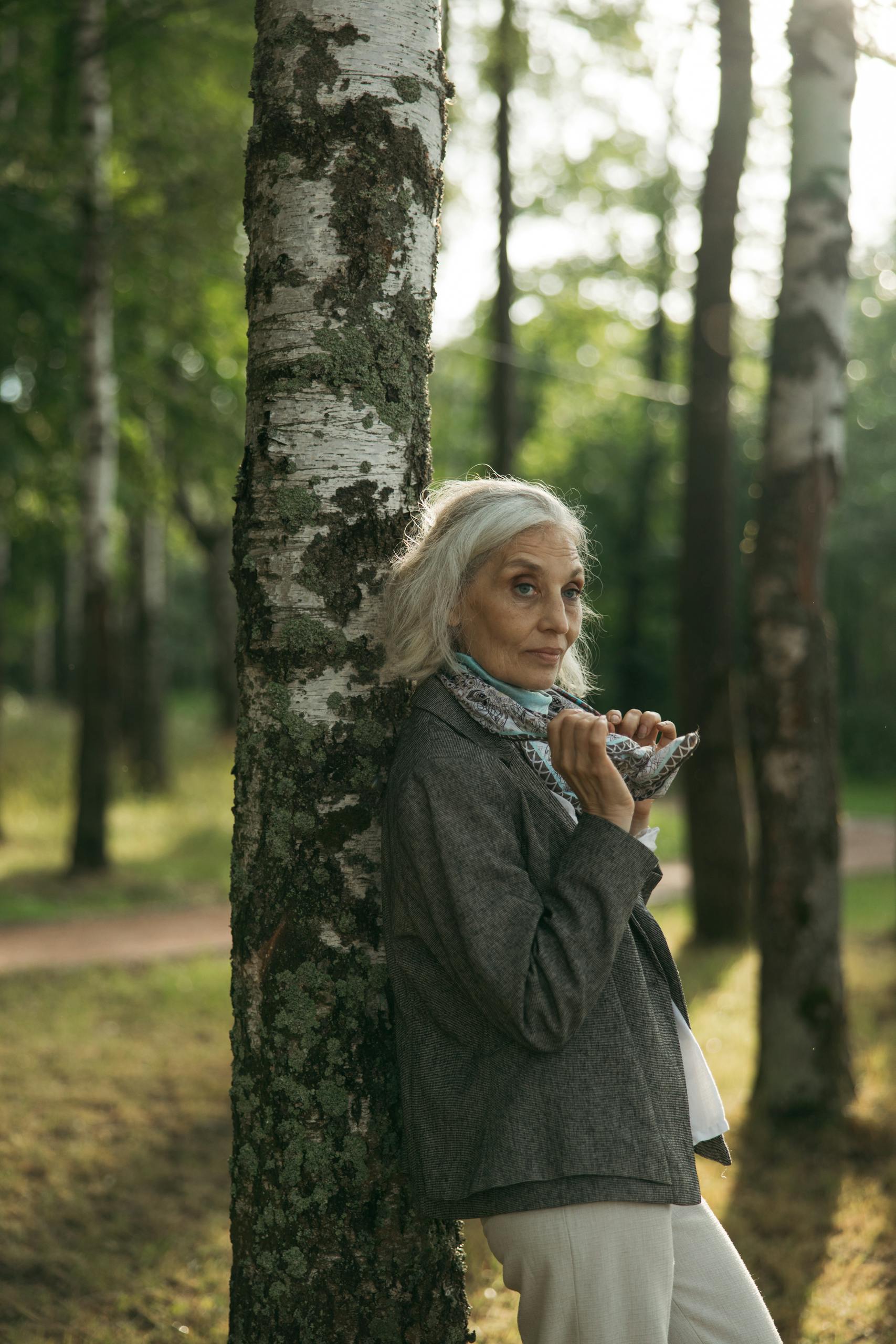 A senior woman leans against a birch tree, surrounded by a serene forest landscape.