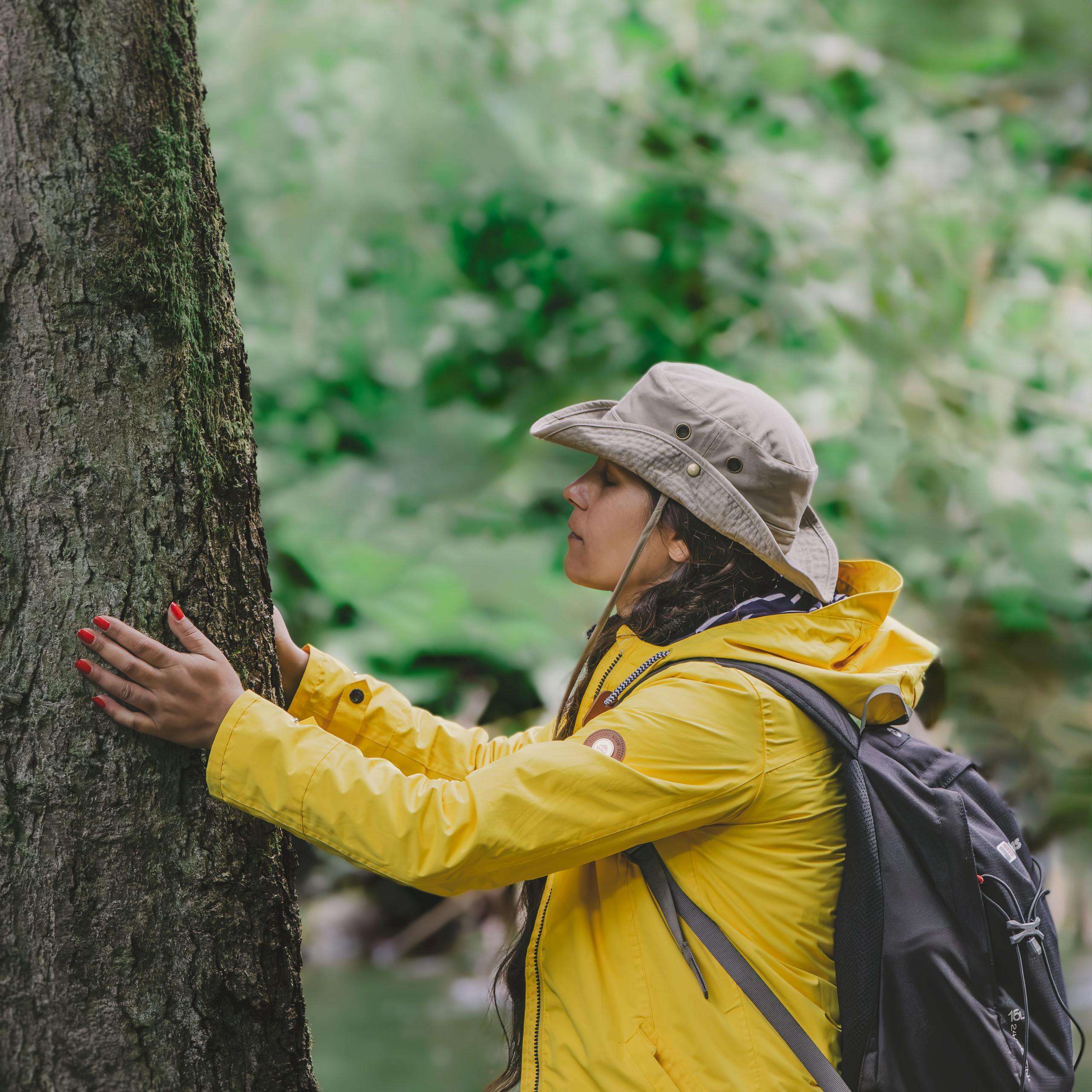 A woman in a yellow jacket embraces a tree in a lush forest, showcasing nature connection.