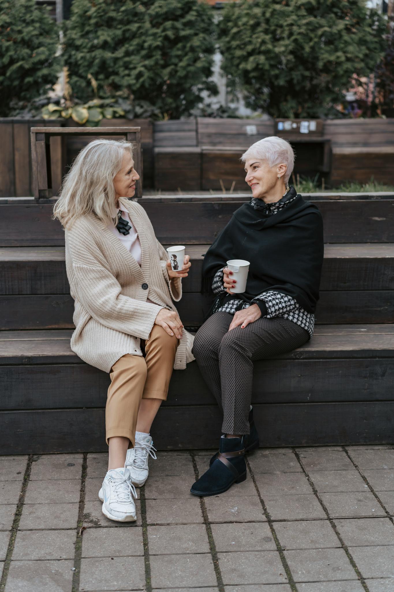 Zwei Frauen sitzen auf einer Treppe im Freien und trinken Kaffee zusammen.