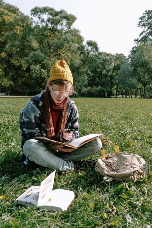 Ein junger Menschn liest entspannt ein Buch im Freien auf einer Wiese; im Hintergrund große Laubbäume, im Vordergrund ein weiteres aufgeschlagenes Buch.