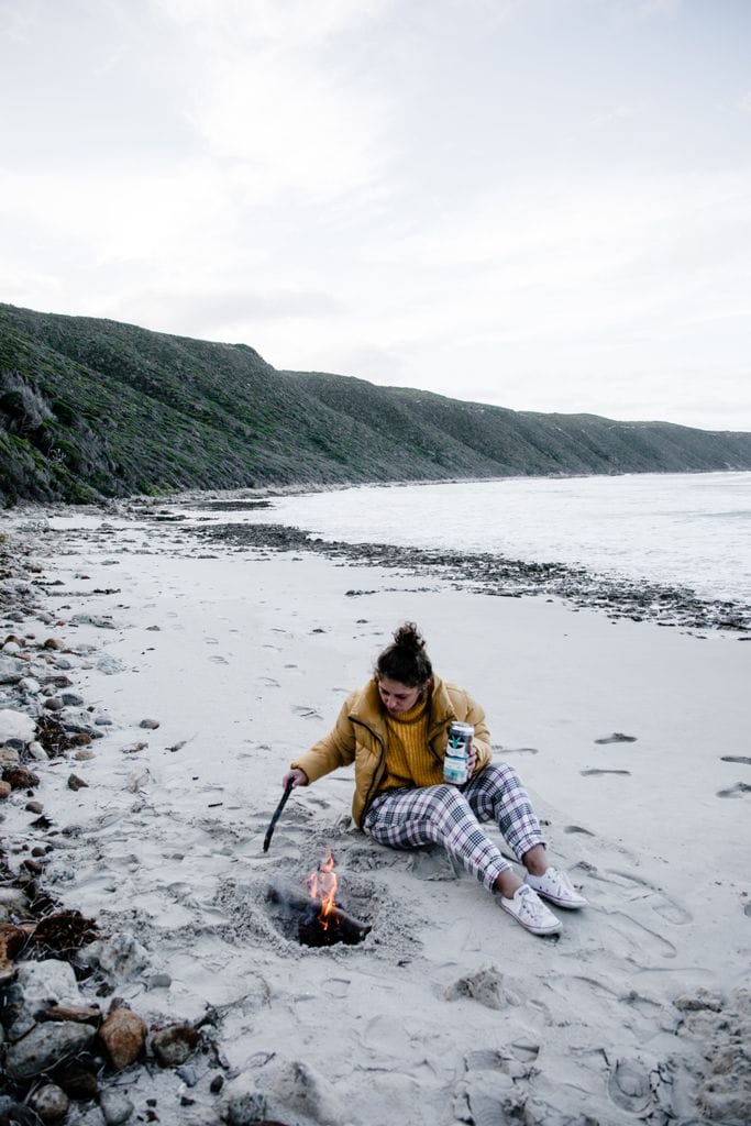 Full length of young female in warm jacket sitting on sandy seashore and warming nea campfire