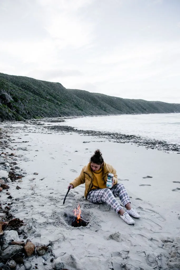 Full length of young female in warm jacket sitting on sandy seashore and warming nea campfire