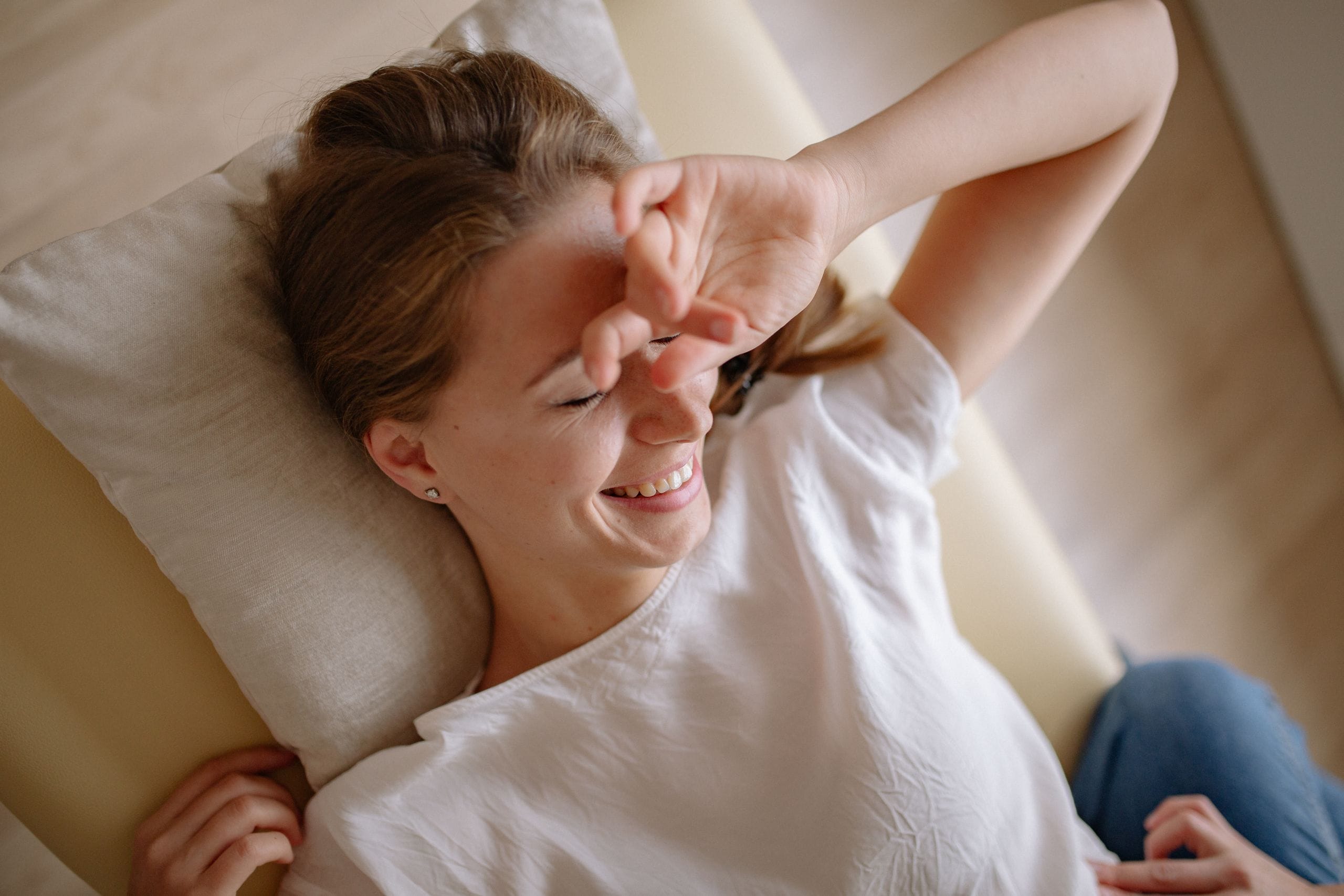 Smiling woman in white top relaxing on bed with hand over face, enjoying leisure time.