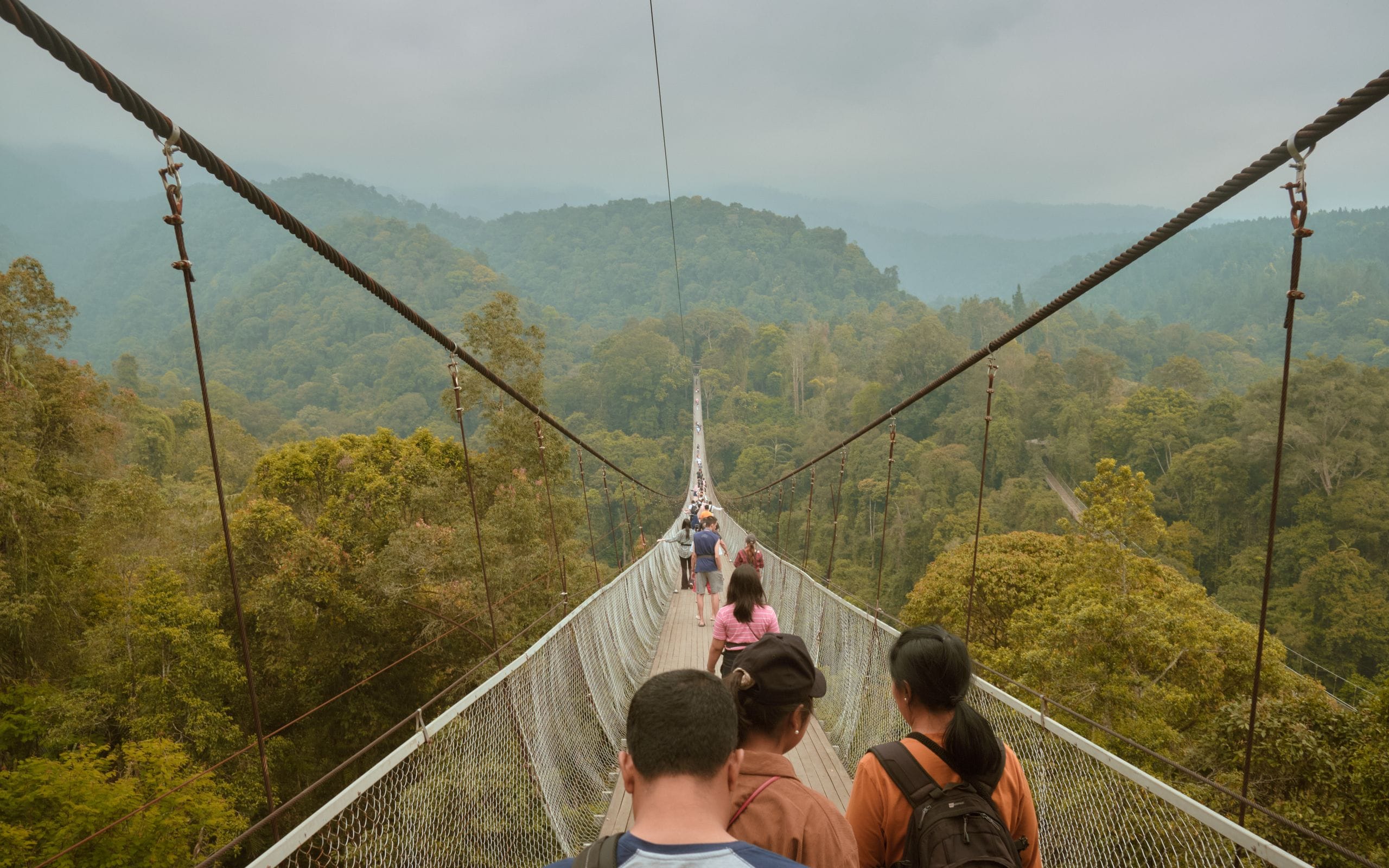 People walking across a large suspension bridge amidst lush greenery in West Java, Indonesia.